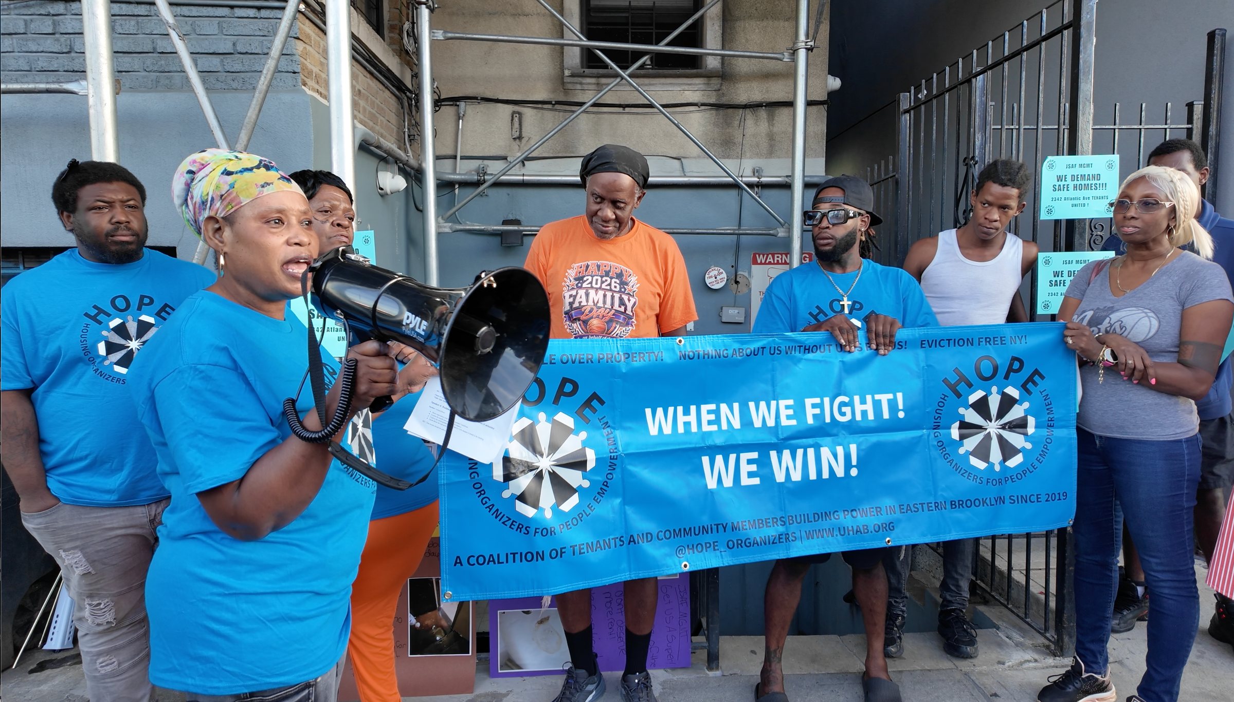Tenants and organizers in blue HOPE T-shirts stand outside 2342 Atlantic Avenue behind a banner reading “When we fight! We win!” as a speaker addresses the crowd through a bullhorn.