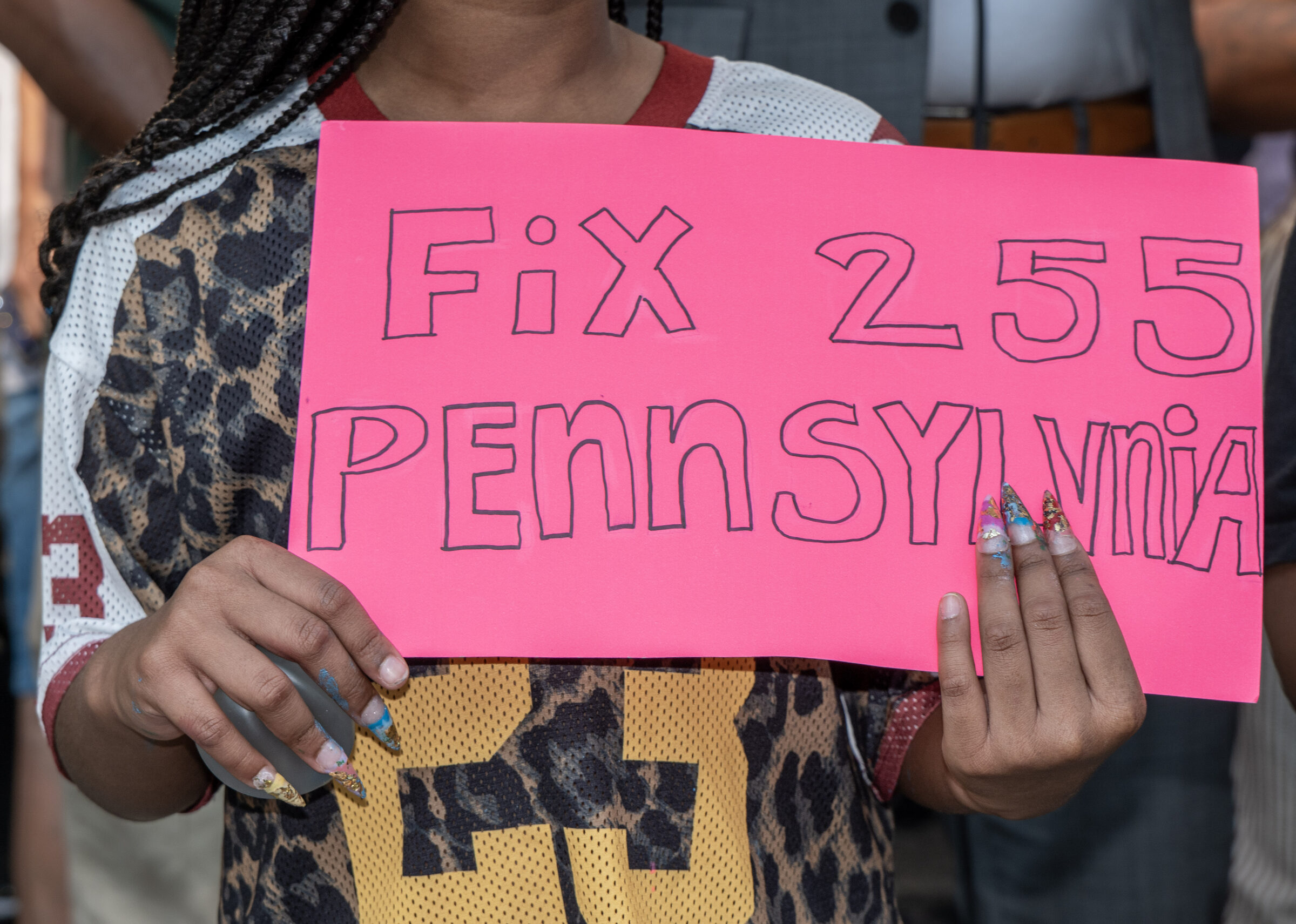 A tenant in a leopard-print mesh jersey holds a hand-lettered pink sign reading &ldquo;Fix 255 Pennsylvania&rdquo; outside the building during the rally.