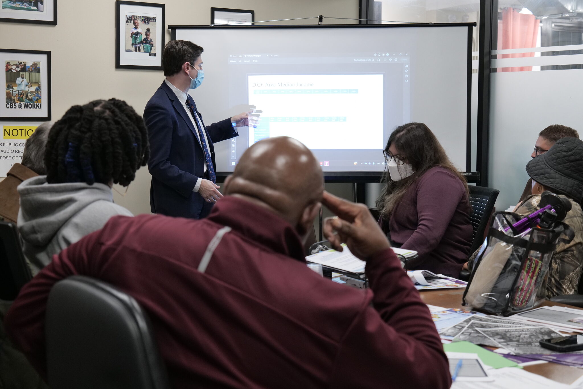A presenter in a suit and face mask points to a screen showing 2026 Area Median Income figures as Community Board 5 committee members and residents look on around a conference table.