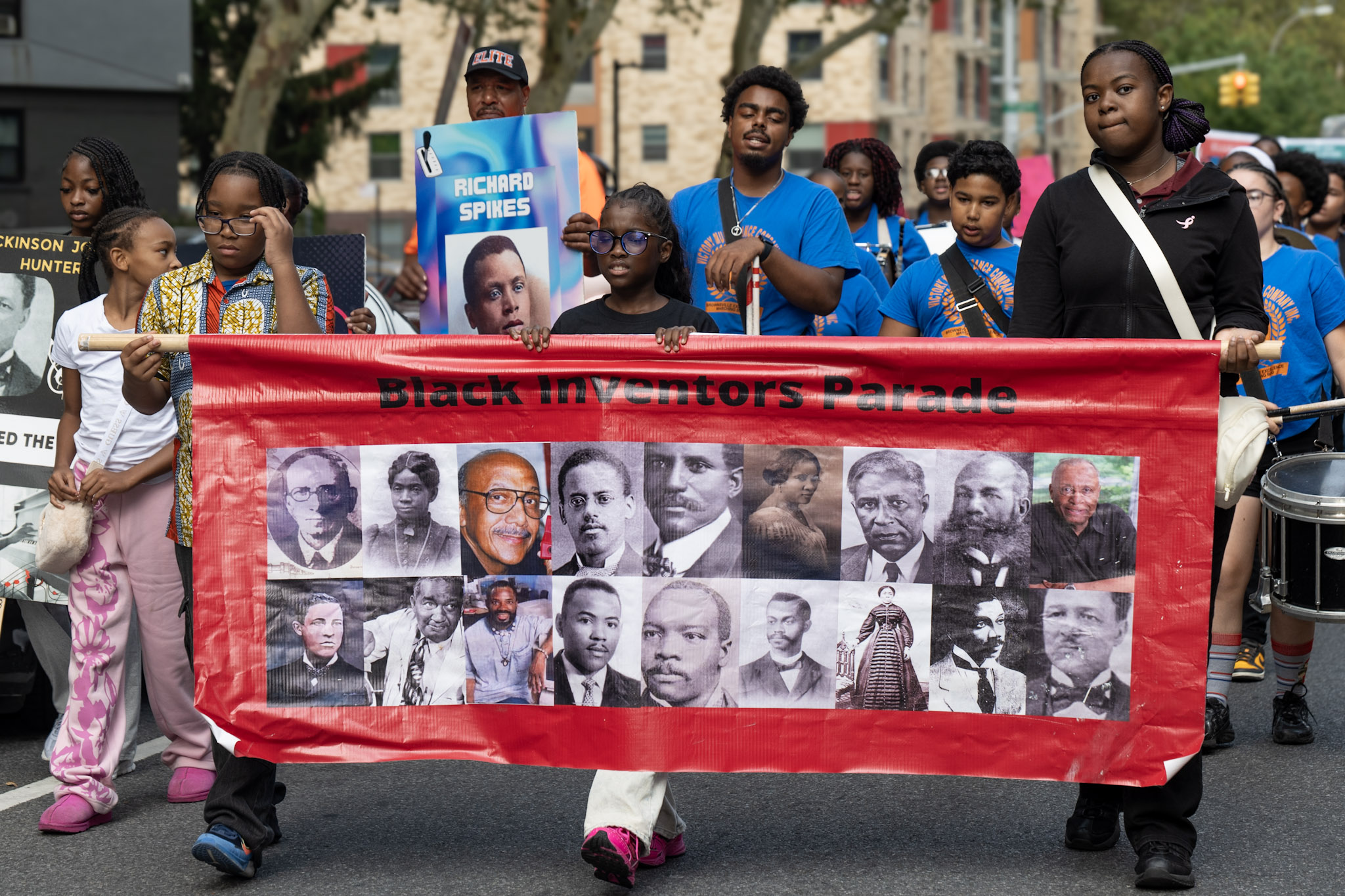 Students march along an East New York street holding a red banner reading “Black Inventors Parade” that is covered with portraits of Black inventors, while a marcher behind them carries a sign for inventor Richard Spikes.