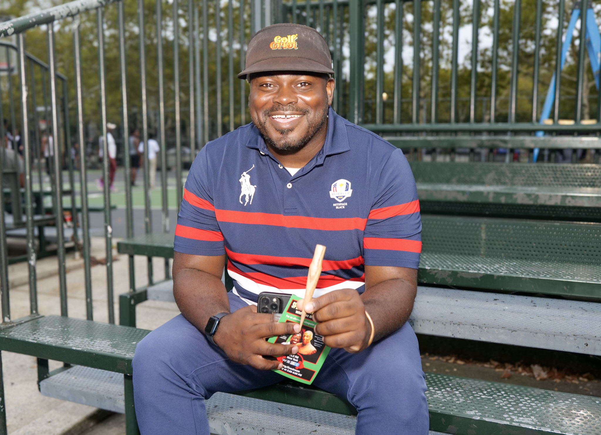Kelley Pierre of All Access Golf LLC sits on metal bleachers in a striped golf polo and visor, holding up a wooden golf tee.