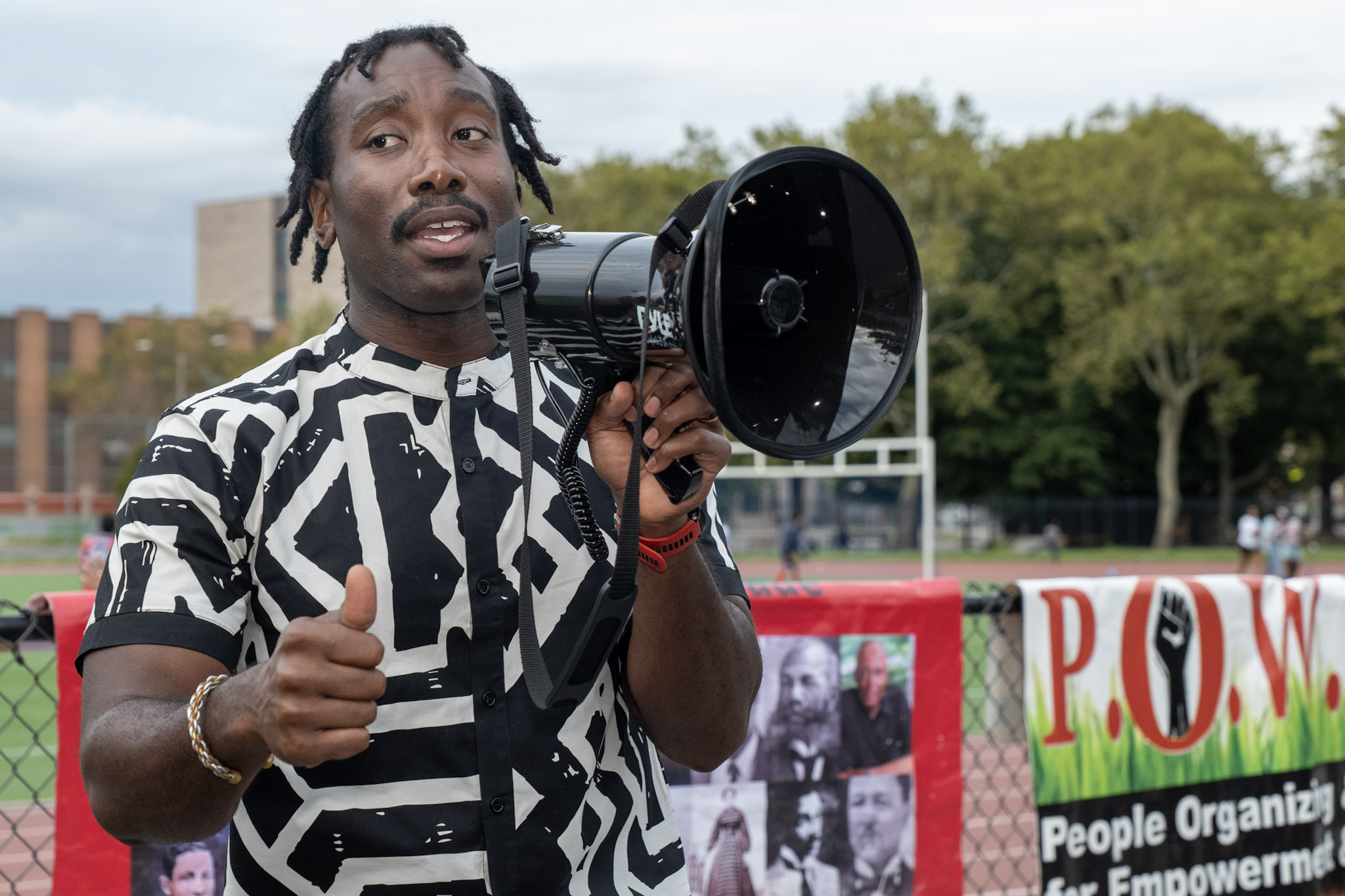 Organizer Keron Alleyne speaks into a megaphone while giving a thumbs up, standing in front of the Black Inventors Parade banner and a People Organizing for Empowerment banner.