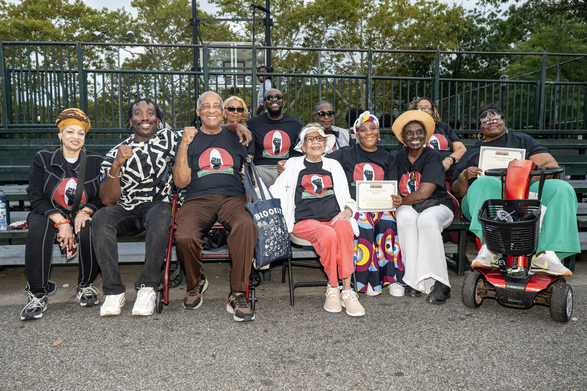 Organizers and community members sit together on bleachers wearing black Operation POWER T-shirts, several of them raising a fist and holding certificates of appreciation.