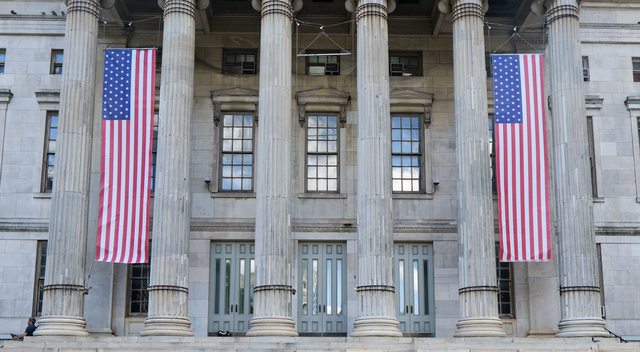 The columned front of Brooklyn Borough Hall in Downtown Brooklyn, with two large American flags hanging between its stone pillars above three sets of doors.