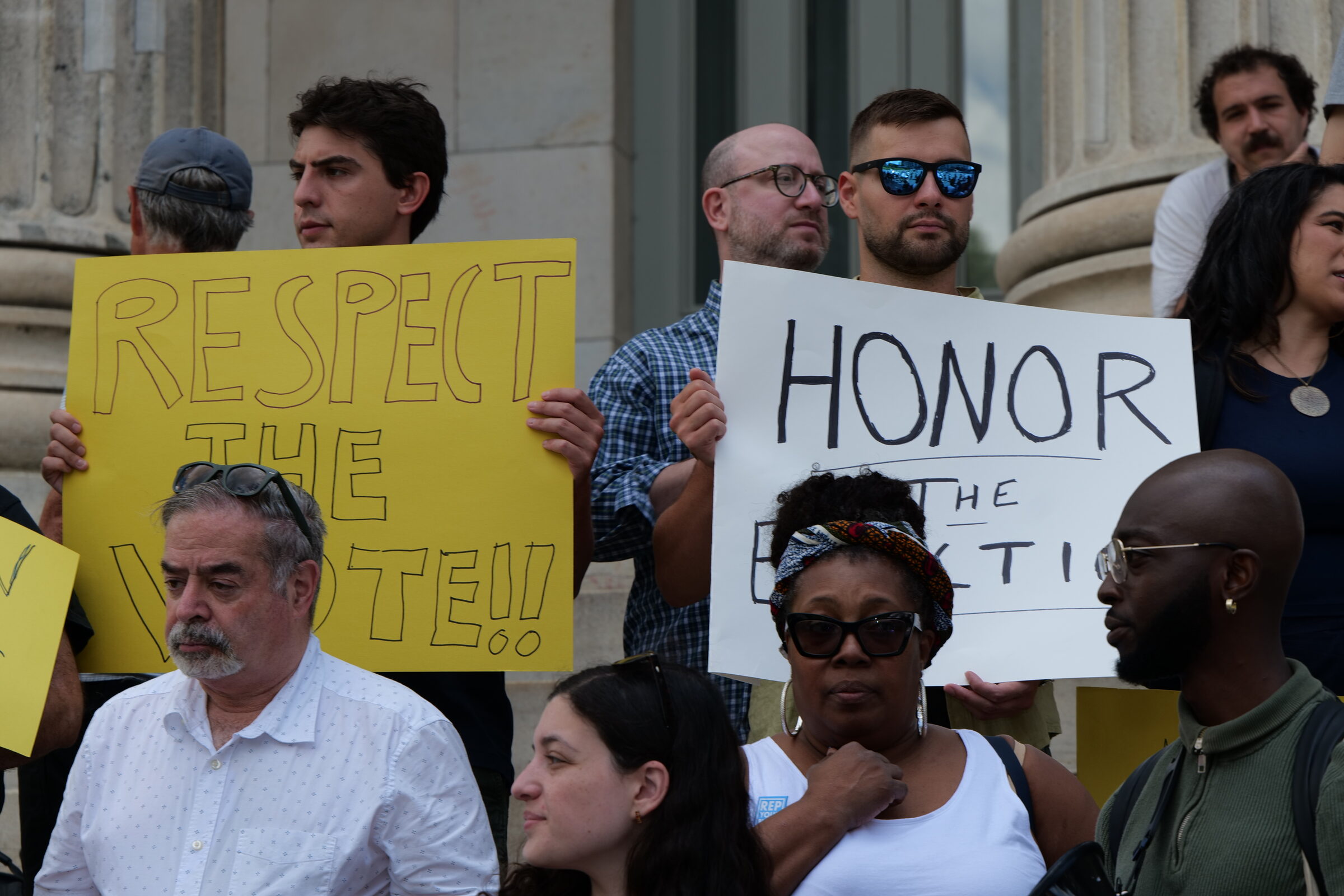 Demonstrators on the steps of Brooklyn Borough Hall hold two hand-lettered signs, one yellow reading &ldquo;Respect the Vote!!&rdquo; and one white reading &ldquo;Honor the Election.&rdquo; Several people stand in the foreground and behind the sign-holders.