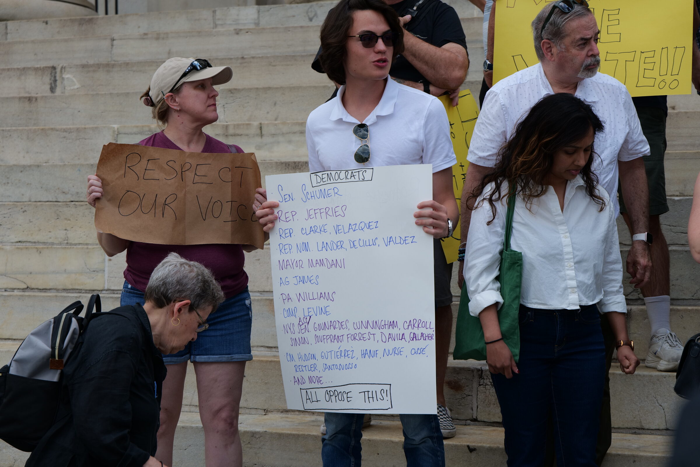 A young man in sunglasses holds a hand-lettered sign on the steps of Brooklyn Borough Hall. The sign is titled “Democrats” and lists names including Sen. Schumer, Rep. Jeffries, Rep. Clarke, Rep. Velázquez, AG James, PA Williams, and Comp. Levine, ending “All Oppose This!”