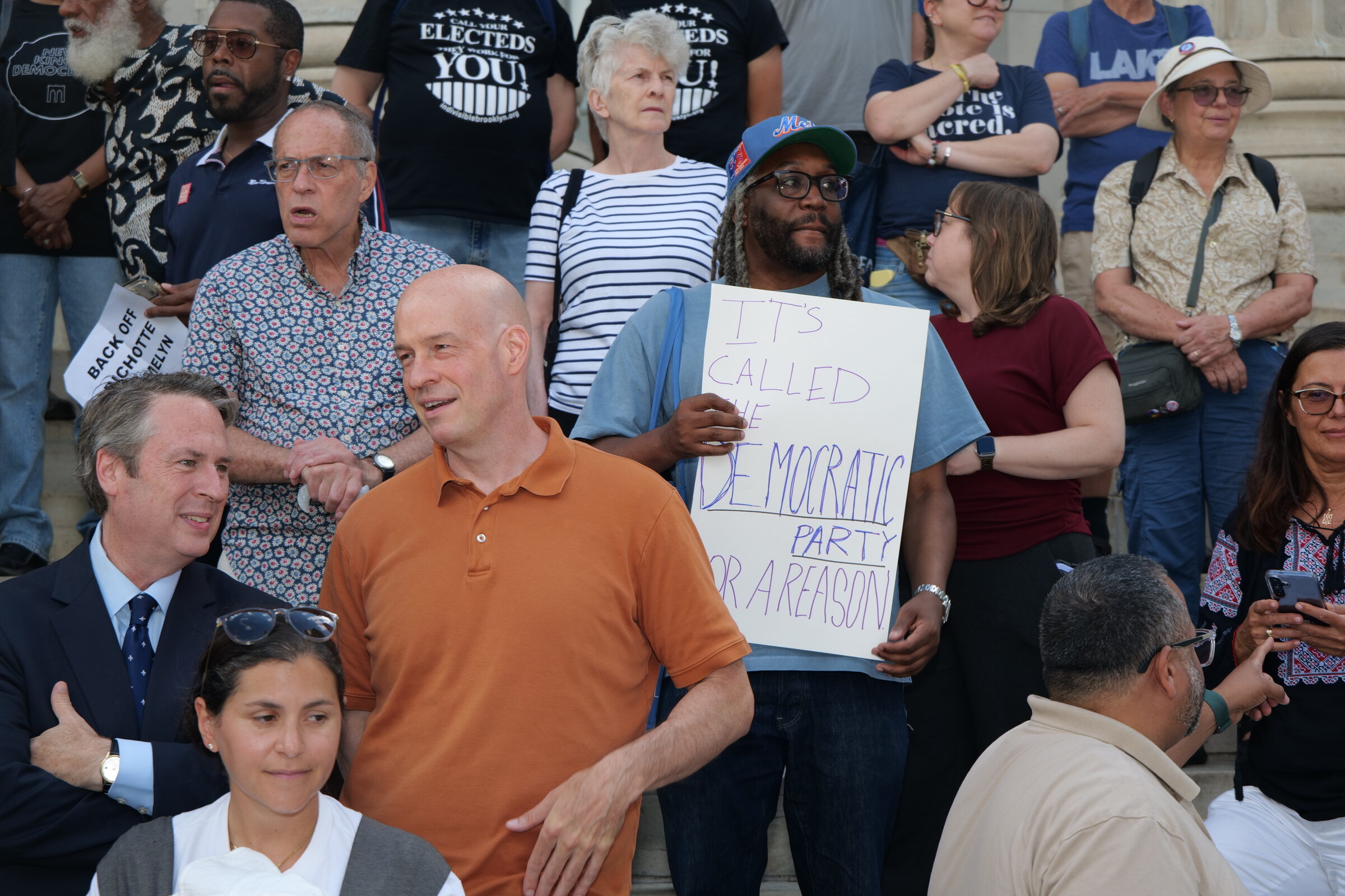 Supporters stand on the steps of Brooklyn Borough Hall at the Brooklyn Can&rsquo;t Wait rally, one holding a hand-lettered sign reading &ldquo;It&rsquo;s called the Democratic Party for a reason&rdquo; and another reading &ldquo;Back off Bichotte Hermelyn.&rdquo;