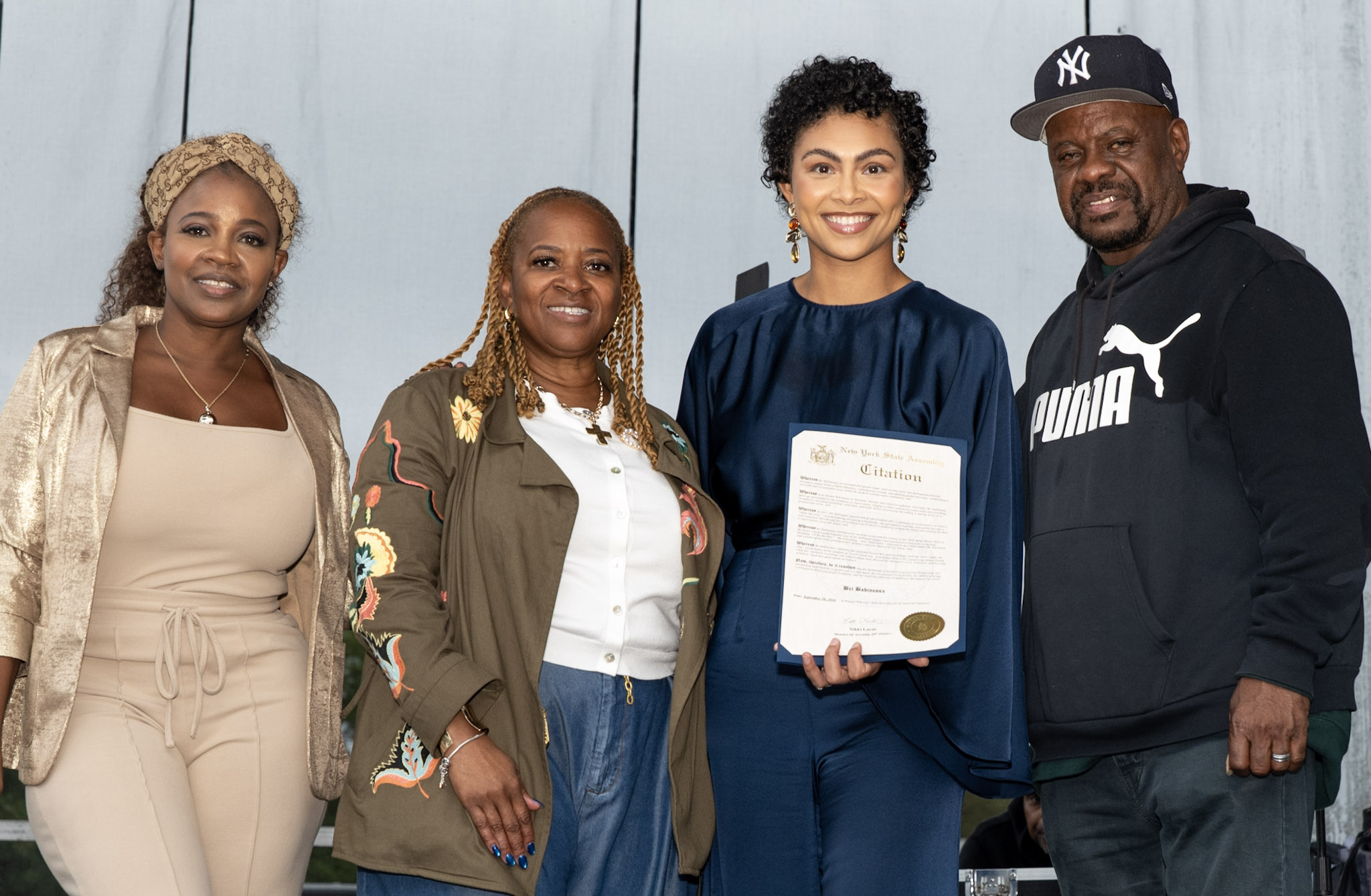 Assemblywoman Nikki Lucas and DJ Keith Watkins stand with gospel artist Bri Babineaux, who holds a New York State Assembly citation onstage.