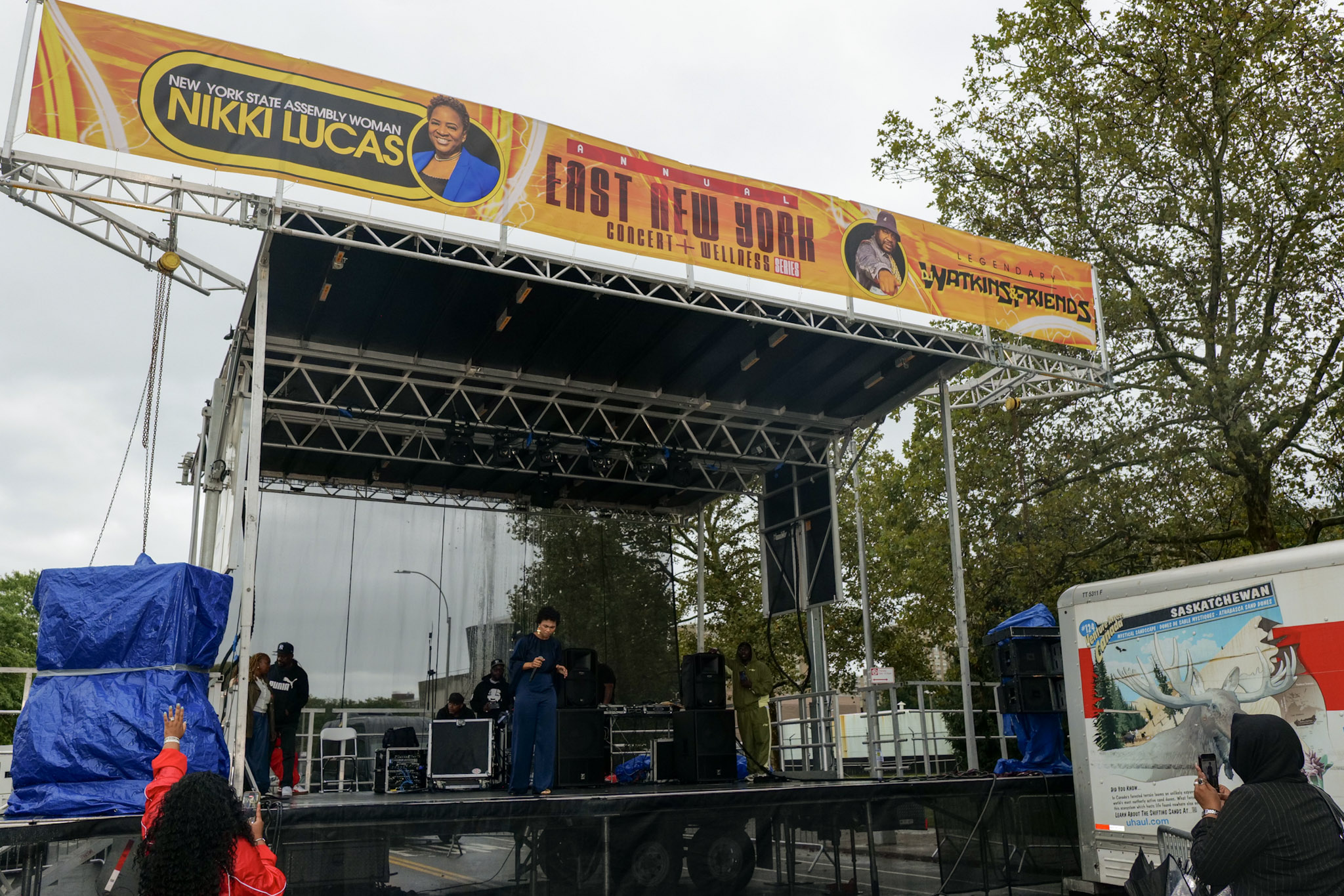 The event stage, topped by a banner for Assemblywoman Nikki Lucas and DJ Watkins & Friends, stands in the rain with covered audio equipment.
