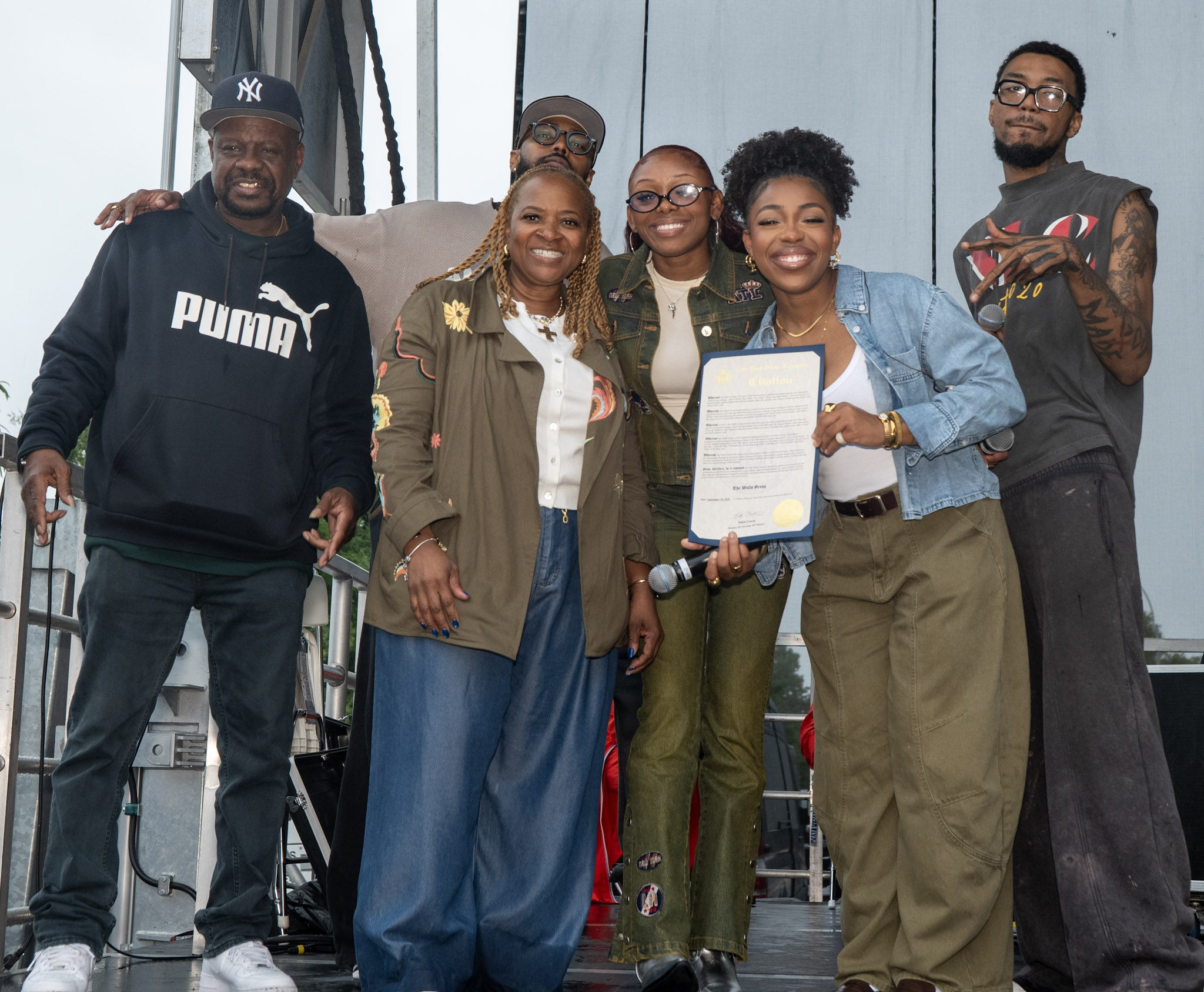 Assemblywoman Nikki Lucas and DJ Keith Watkins stand onstage with The Walls Group, who hold a New York State Assembly citation.