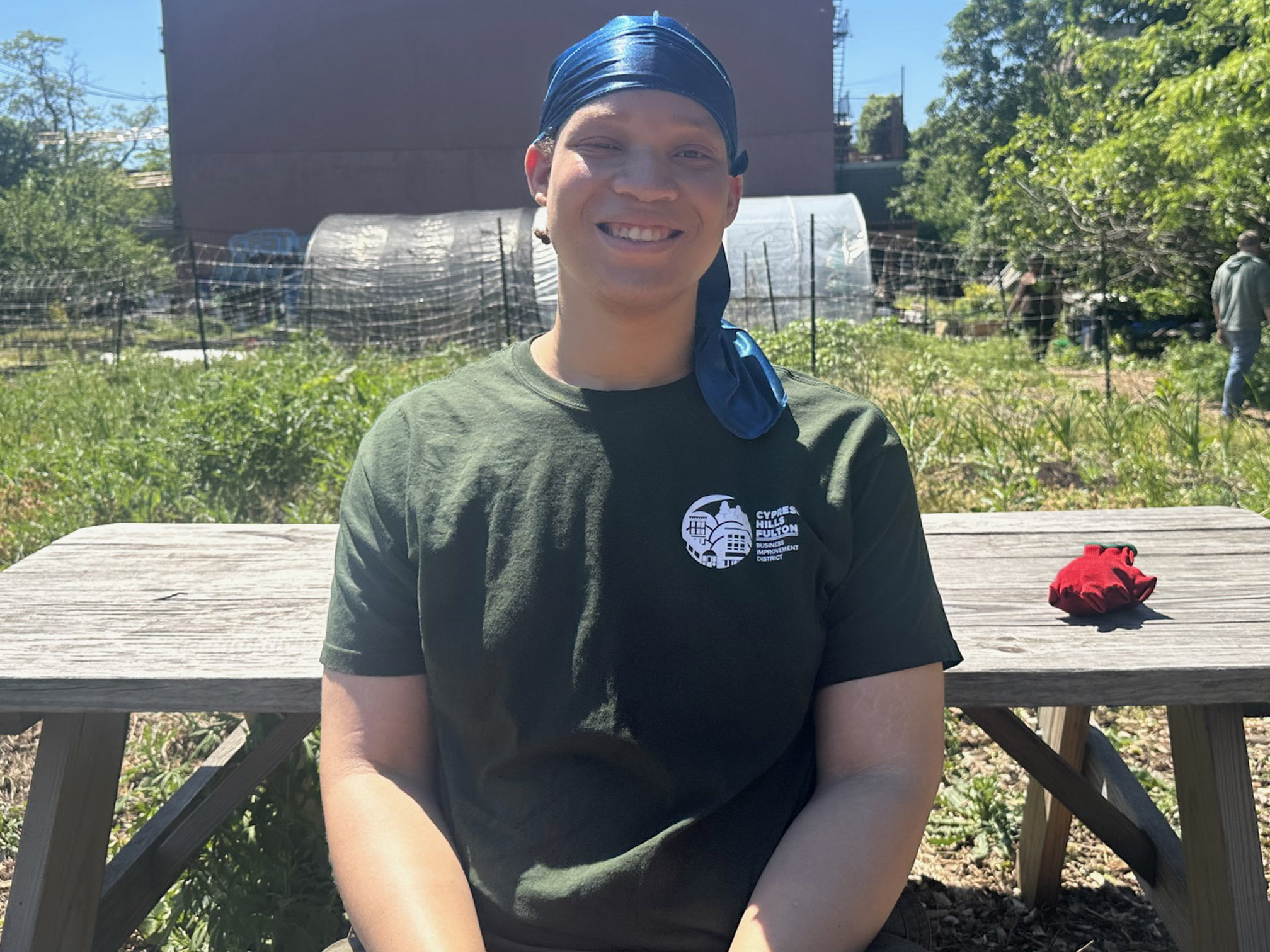 Joshua John, smiling in a blue durag and a dark green T-shirt sits at a wooden picnic table on the farm, with garden beds, hoop houses and a brick building behind him.