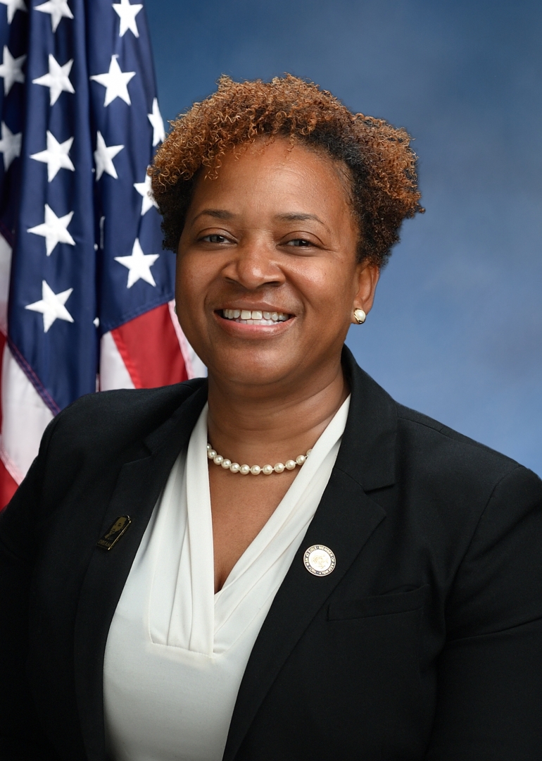 Official portrait of Assemblywoman Nikki Lucas, smiling, in a black blazer and pearl necklace in front of an American flag.