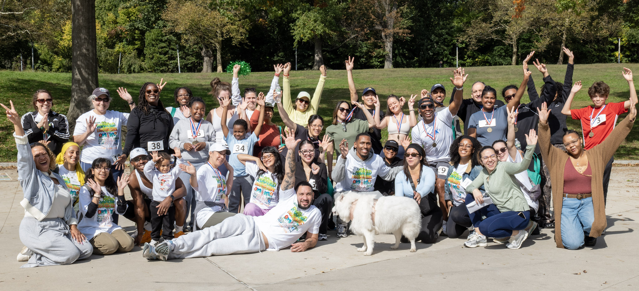 Dozens of runners, volunteers and families, many wearing medals and colorful Rooted in East New York T-shirts, wave and raise their arms for a group photo on a sunny plaza in Highland Park, with a fluffy white dog in front.